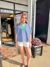 Load image into Gallery viewer, Woman in a blue checkered top and white shorts standing outside a store with flowers and a sign in the background.