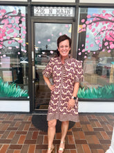 Load image into Gallery viewer, A woman stands smiling in front of a storefront window wearing a short-sleeve patterned dress with a high collar and ruffled trim. The dress has a brown, beige, and black wavy abstract print.