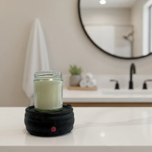 Load image into Gallery viewer, A glass jar candle filled with pale cream wax sits on a black circular electric warmer with a small red power button, setting on a bathroom counter.