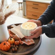 Load image into Gallery viewer, A person holds a three-wick candle in a white pumpkin-shaped ceramic holder, with two lit flames visible. The scene is styled on a dark wooden tray with decorative pumpkins and a neutral home interior in the background.