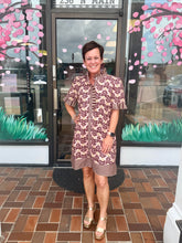 Load image into Gallery viewer, A woman stands smiling in front of a storefront window wearing a short-sleeve button-front dress with a brown and beige wavy abstract pattern and ruffled sleeves. The storefront has pink cherry blossom decals and a brick floor.